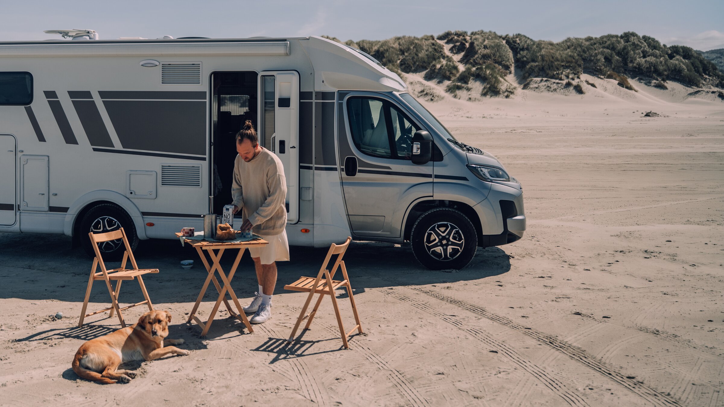 Carado Wohnmobil am Strand, Person bereitet Frühstück am Campingtisch zu, Hund liegt davor im Sand