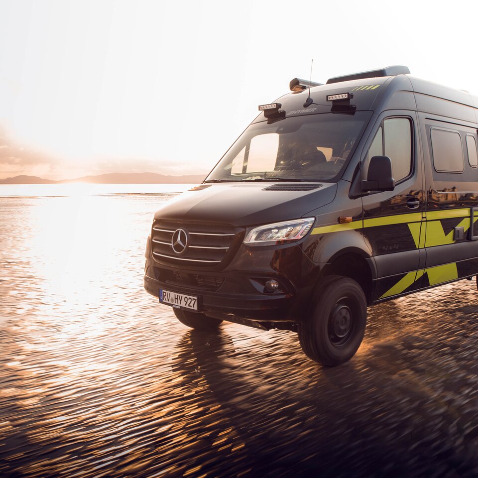 Le camping-car tout-terrain Mercedes HYMER Grand Canyon S roule rapidement sur une plage ensoleillée et humide. L'environnement est flou en raison de la vitesse.