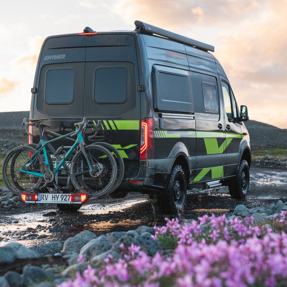 Le camping-car tout-terrain Mercedes Sprinter HYMER Grand Canyon S CrossOver est stationné sur un chemin non aménagé avec des fleurs et de l'herbe, des vélos sont sur le porte-vélos.