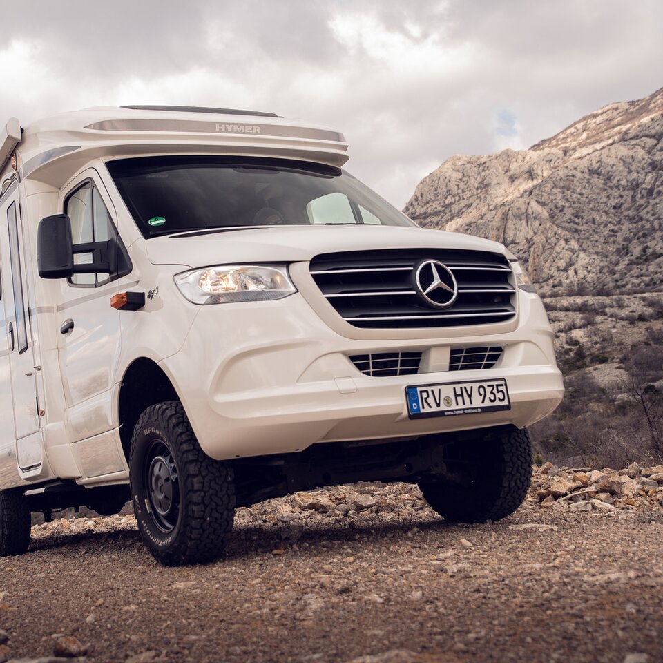 Le camping-car tout-terrain Mercedes tout-terrain HYMER ML-T blanc est stationné sur une route de montagne avec des collines rocheuses et un ciel légèrement nuageux.