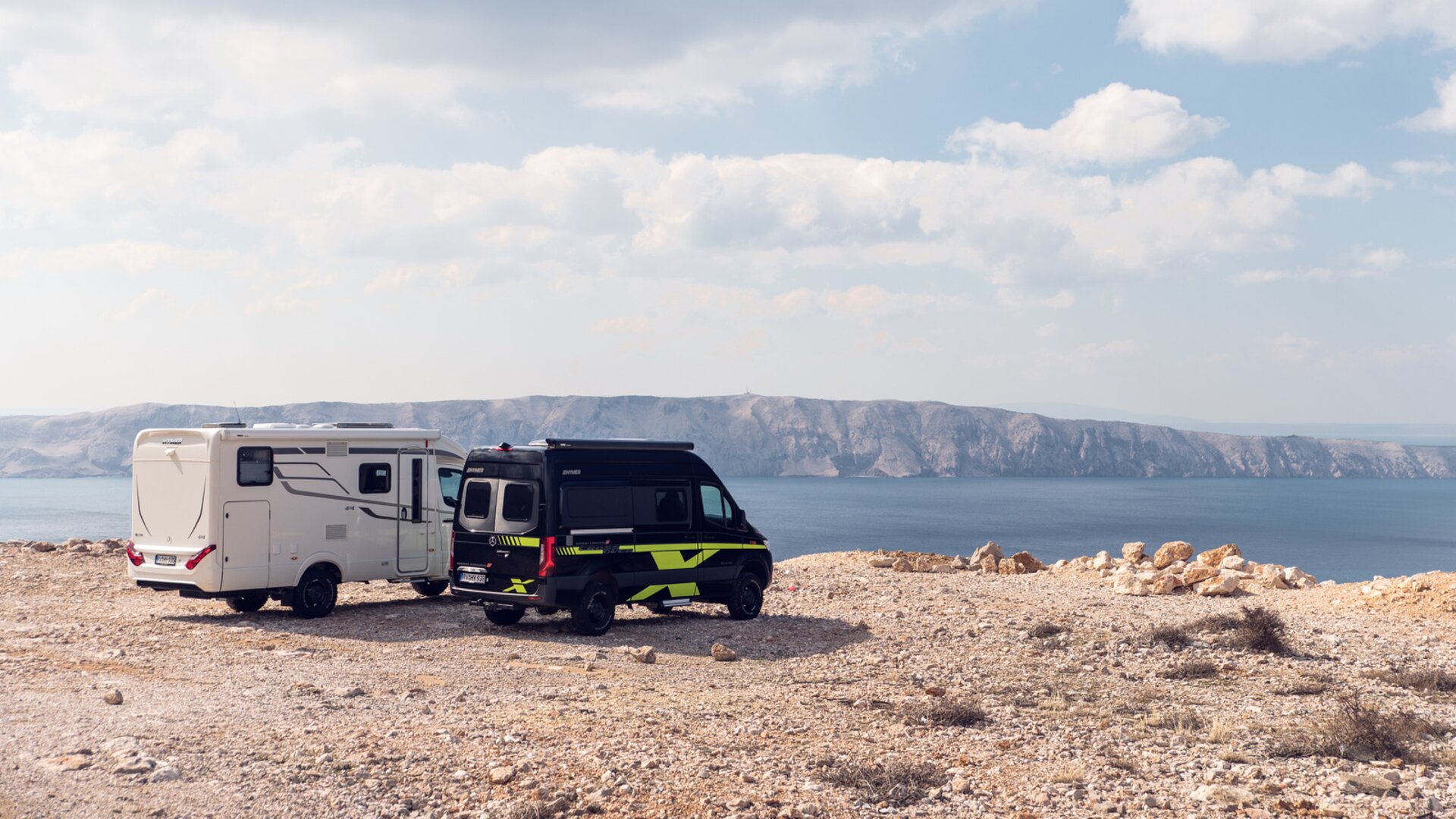 Deux camping-cars tout-terrain blanc et noir-vert sont stationnés sur une plaine ensoleillée surplombant la mer, des falaises sont visibles à l'arrière-plan.