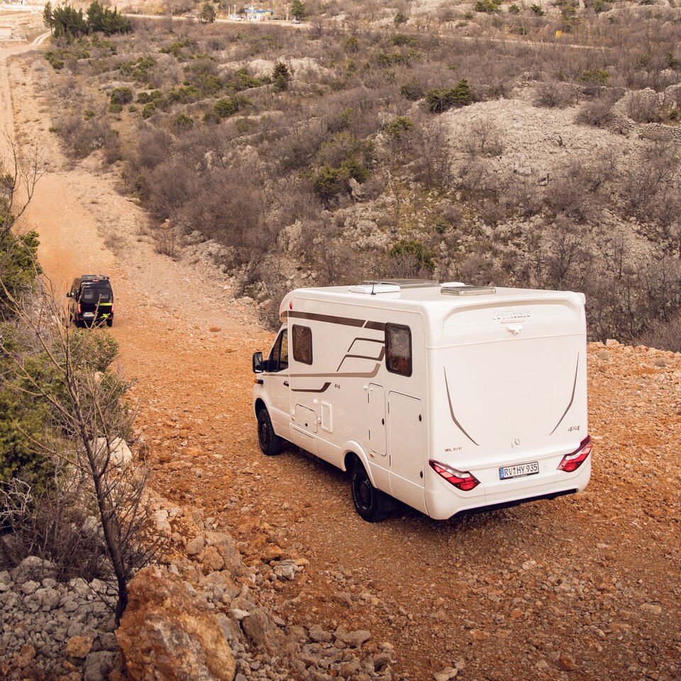 Deux camping-cars tout-terrain 4x4 de HYMER avec tête motrice Mercedes roulent sur une route vallonnée à travers un terrain non aménagé et rocailleux avec des buissons.