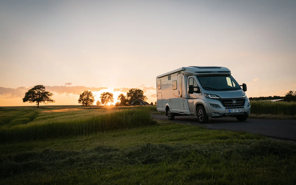 HYMER Exsis-t på en smal vei midt i grønne enger og kornåkre ved solnedgang