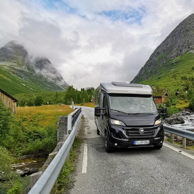 HYMER Exsis-t på en smal fjellvei omgitt av steinete fjell, grantrær, grønne enger og fjellbekk