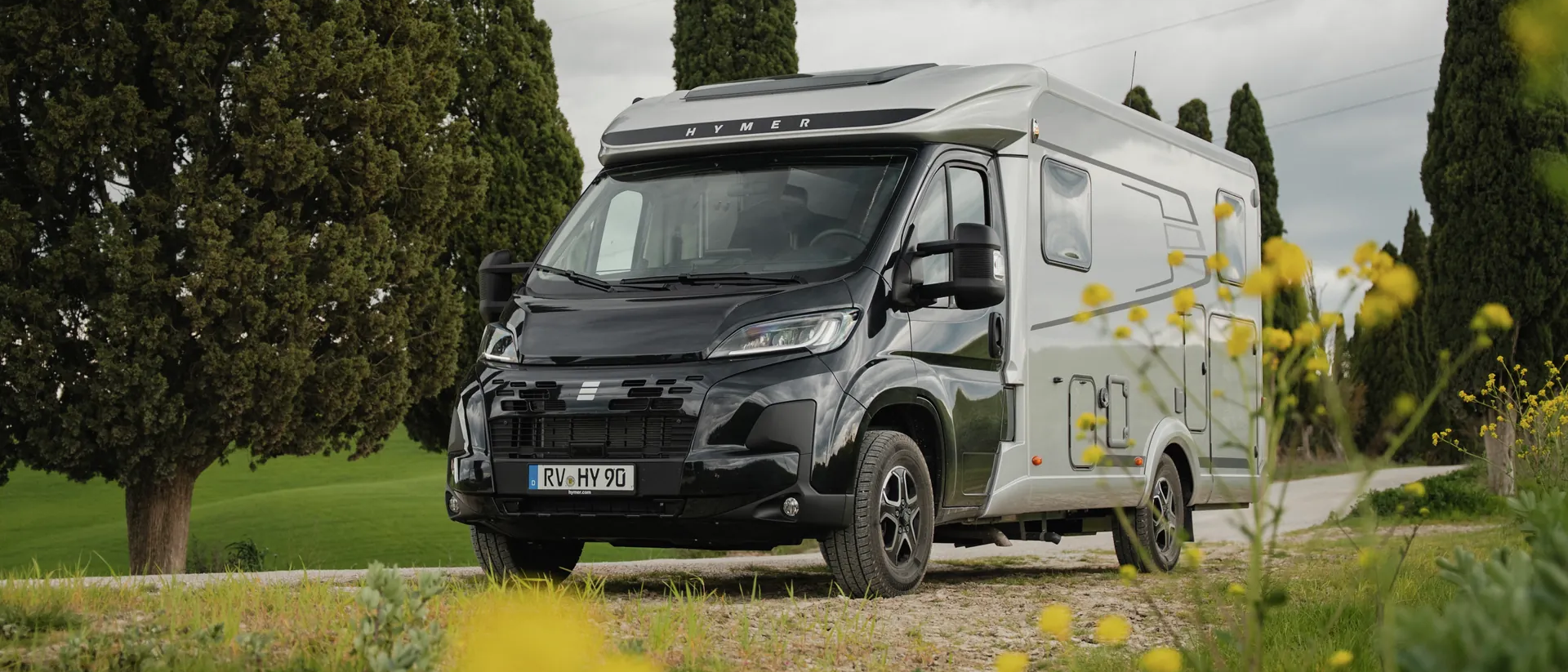 Hymer Exsis‑t med svart førerhus og sølvfarget bodel står på en landevei i et landlig landskap med sypresser, blomster i forgrunnen og overskyet himmel.