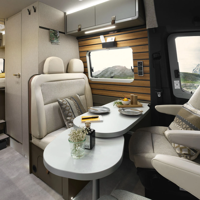 Modern interior of a campervan with light upholstery, showing the seating area with a swivelled-out living room table. The wall at the seating area is clad with wood panels.