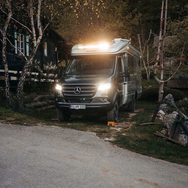 Semi-integrated motorhome seen from the front and side, standing at dusk next to a wooden hut in a small forest, headlights and LED light bar are on